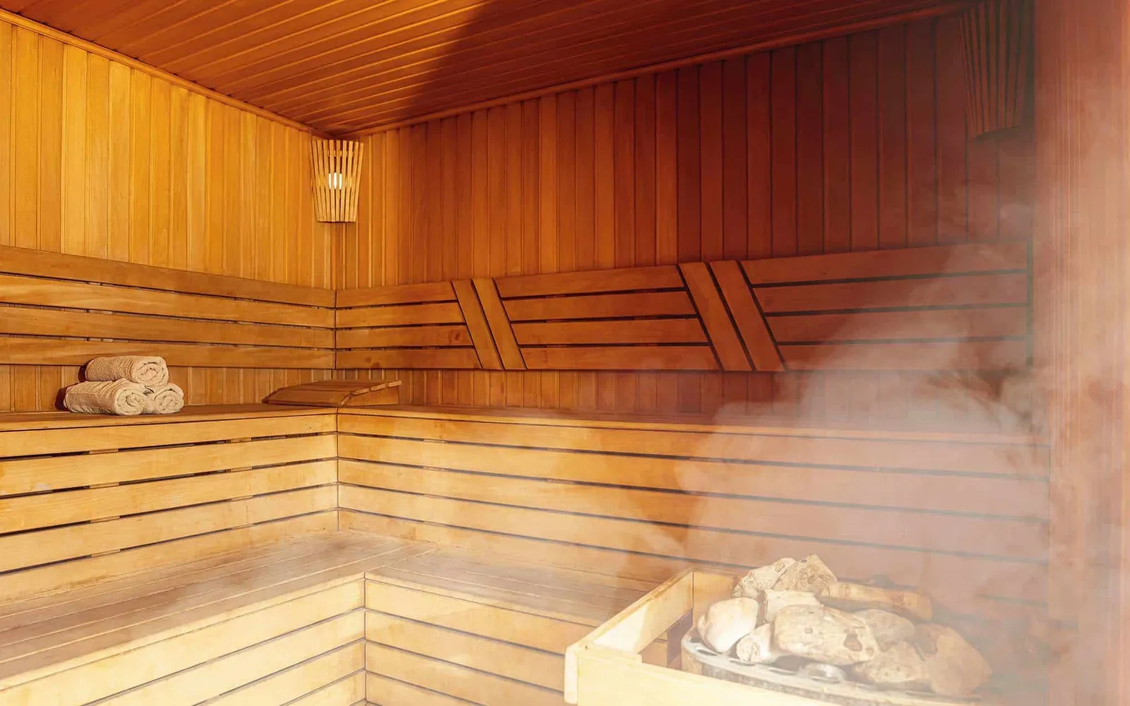 A wood-panelled sauna interior with tiered benches, rolled towels and a stone heater releasing steam