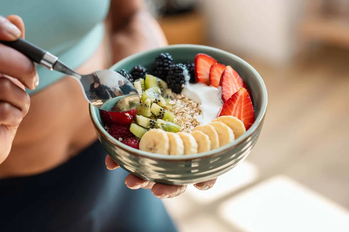 A person holding a bowl of oats topped with banana, strawberries, blackberries and kiwi
