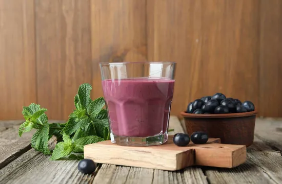 A glass of purple berry smoothie beside fresh mint leaves and a bowl of blueberries on a wooden board