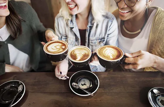 A group of women laughing together while clinking cups of latte art coffee at a wooden cafe table