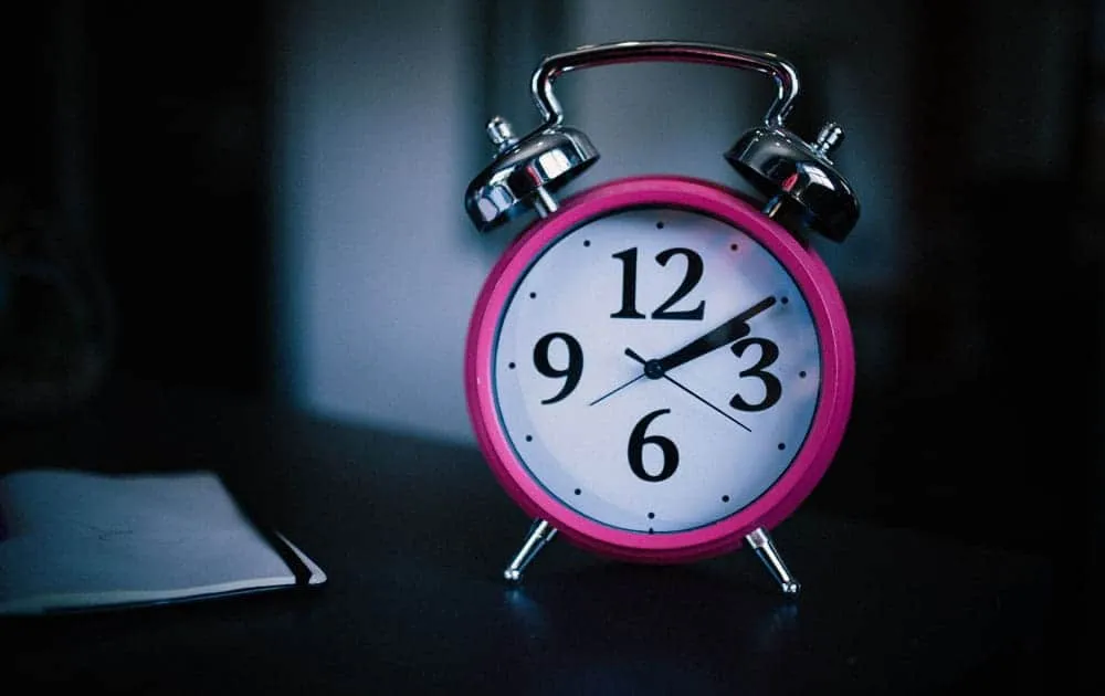 A vintage-style pink alarm clock sits on a dark bedside table beside a notebook