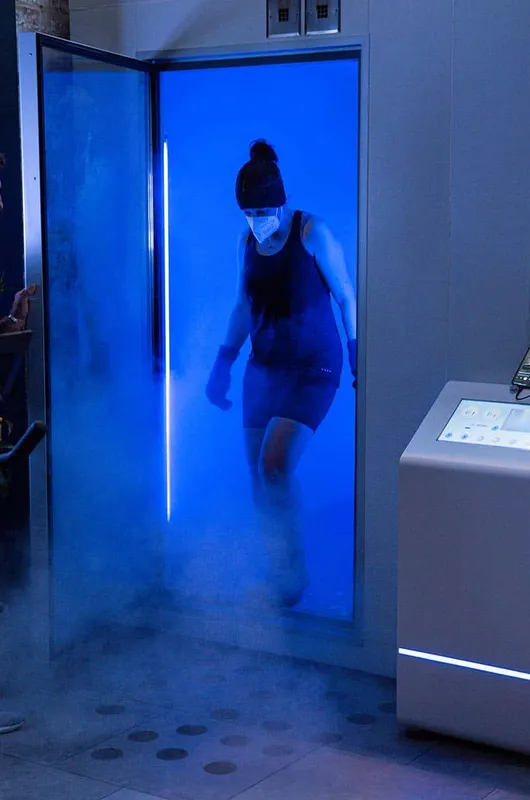 Close-up profile portrait of a woman wearing a beanie and face mask lit by blue light from a cryotherapy chamber