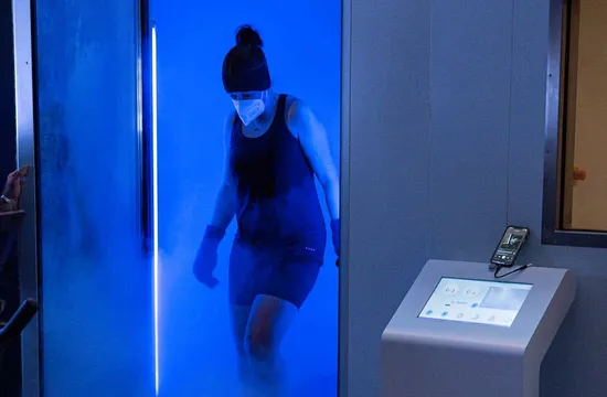 A woman in a beanie, mask and gloves emerging from a cryotherapy chamber surrounded by cold blue light and mist