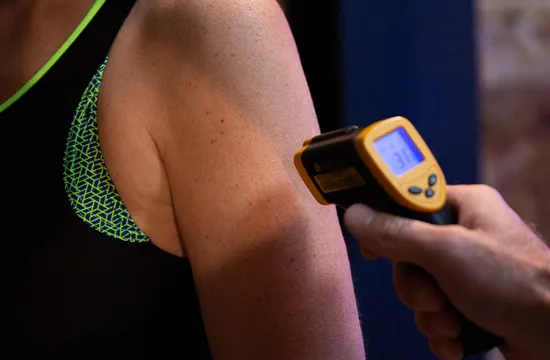 A hand holding a yellow infrared thermometer to measure skin temperature on someone's upper arm