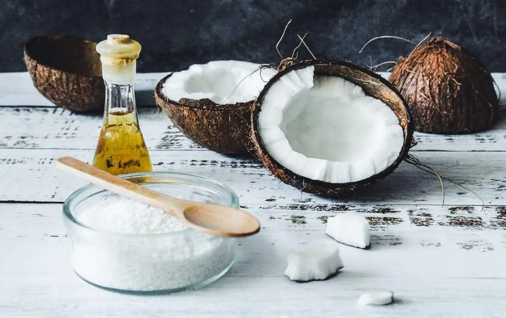 Cracked coconut halves sit beside a jar of coconut oil and a wooden spoon on a table