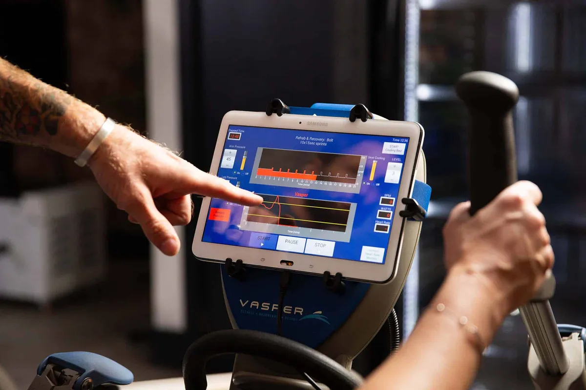 A tattooed hand points at a Vasper machine's touchscreen displaying workout metrics