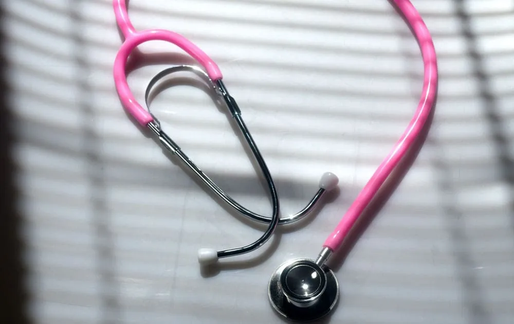 Pink stethoscope resting on a white surface, softly lit by window blinds