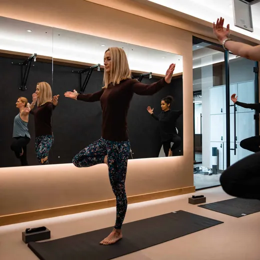 A woman balances in tree pose with arms outstretched on a yoga mat, reflected in the studio mirror behind her